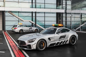 兩輛銀色 Mercedes-AMG F1 安全車，閃著燈，停在一棟現代玻璃建築前。 — MacBook Pro 14 壁紙