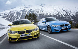 兩輛BMW跑車在白雪覆蓋的山峰背景下，於山路上飛馳。