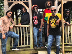 六位雷鬼風格的男士在木質門廊上戴著太陽眼鏡和彩色襯衫擺姿勢。 — 巴布·馬利語錄 壁紙