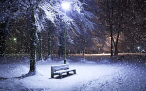 積雪覆蓋的長凳在冬日公園中，被街燈照亮，雪花飄落。 — 冬季風景 壁紙