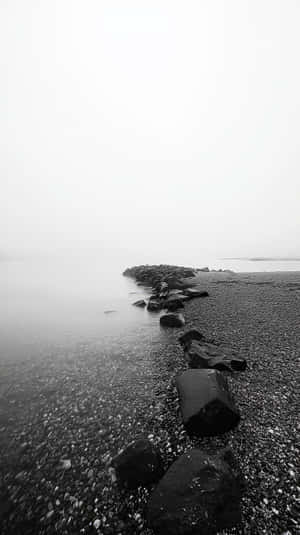 黑白海灘，岩石和鵝卵石在陰天天空下延伸至霧氣瀰漫的水面。 — 抽象 壁紙