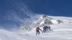 四位登山者在有風的條件下走在積雪的山坡上。 — 雪山 壁紙
