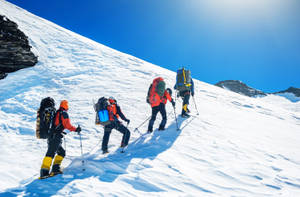 四名登山者穿著鮮豔裝備在藍天下攀登雪山斜坡。 — 攀爬 壁紙