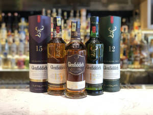 四瓶 Glenfiddich 威士忌和盒子放在大理石吧台上，後面有模糊的酒架。 — 酒吧 壁紙