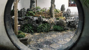圓形石窗框住一個寧靜的傳統中式花園，內有岩石、竹子和馬賽克小徑。 — 16:9 壁紙