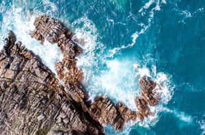 在明亮的日光下，崎嶇的海岸岩石被碧綠色的海浪拍打。 — 海灘 壁紙