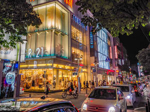 夜晚的城市街道，有發光的商店、汽車和人們在樹下行走。 — 永遠21 壁紙