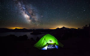 在星空下夜間搭建的發光綠色帳篷，湖泊和山脈上方可見銀河。 — 山脈 壁紙