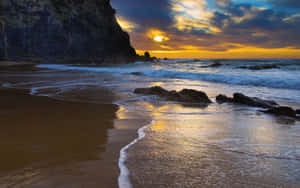 夕陽下的岩石海灘，海浪拍打海岸，懸崖在背景中 — 海灘日落 壁紙