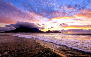 夕陽下的桌山，海浪在多彩雲層下拍打沙灘。 — 開普敦 壁紙