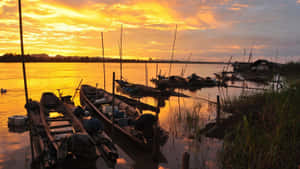 金色夕陽下，木船停泊在河岸邊，附近有浮動房屋。 — 夕陽天空 壁紙