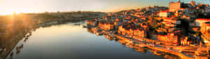 波爾圖歷史悠久的河畔上空金色夕陽，建築物和船隻倒映在平靜的水面上。 — 32:9 壁紙