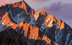 雪山山峰在夕陽下閃耀橙色光芒，伴隨紫色雲彩 — macOS 壁紙