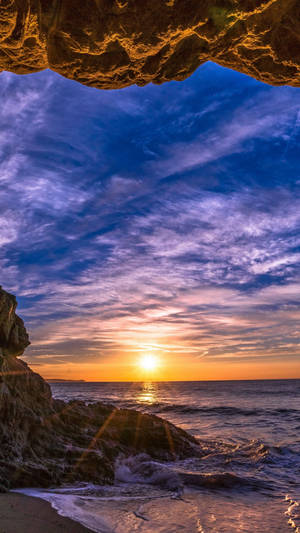 從岩石懸垂下方觀看的夕陽，下方有波浪拍打。 — 日落 壁紙