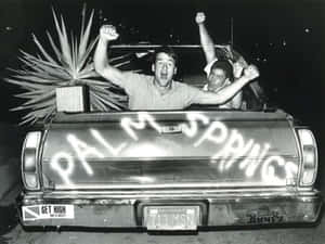 夜晚，兩名男子在尾門上噴有「PALM SPRINGS」的皮卡車中慶祝。 — 70年代動圖 壁紙