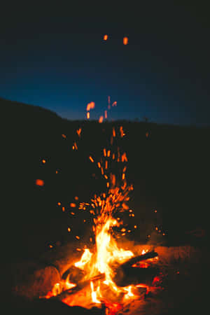 營火火花在夜間向上飛舞，背景是黑暗的山丘。 — 2:3 壁紙