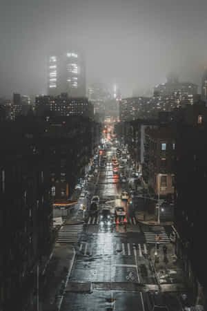 夜晚一條潮濕的城市街道，閃爍的車燈和遠處霧濛濛的摩天大樓。 — 手機 壁紙