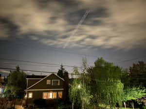 一間窗戶亮燈的房子，夜空中有飛機尾跡和樹木。 — 月夜星空 壁紙