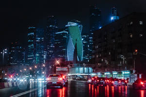 夜晚城市街道，發光的建築物和潮濕的人行道反射汽車燈光。 — 城市燈光 壁紙