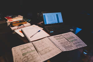 夜晚昏暗燈光下，雜亂的書桌上放著打開的筆記本、筆記型電腦和書籍。 — 讀書 壁紙