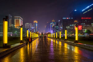 夜晚城市步道，發光的黃色柱子和摩天大樓在潮濕的路面上反射。 — 城市燈光 壁紙