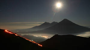 夜晚噴發的火山，發光的熔岩流和上方明亮的月亮。 — 瓜地馬拉 壁紙