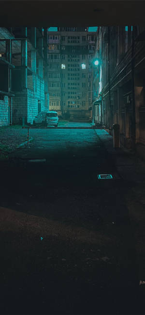 夜晚昏暗的城市小巷，青色光芒下有一輛停泊的貨車和高樓。 — iPhone 15 Plus 壁紙