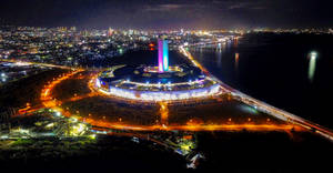 夜晚鳥瞰一座發光的圓形建築靠近水邊，伴隨著城市燈光。 — 城市風景 壁紙