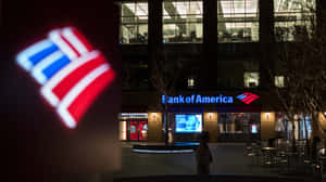 夜間，一個人走過燈火通明的Bank of America大樓，前景有發光的標誌。 — 美國銀行 壁紙
