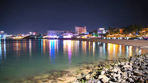 夜間海岸城市，色彩繽紛的燈光映照在岩石海岸附近的水面上。 — 海洋 壁紙