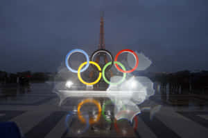 夜間奧運五環亮起，背景為艾菲爾鐵塔，濕漉漉的人行道反射光線。 — MacBook Pro M2 壁紙