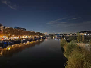夜晚河景，城市燈光倒映在水面上，船隻停泊在樹木成行的河岸邊。 — 秋天 壁紙