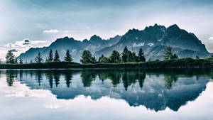 寧靜的山湖在藍天下映照出鋸齒狀的山峰和常綠樹木。 — 冬季風景 壁紙