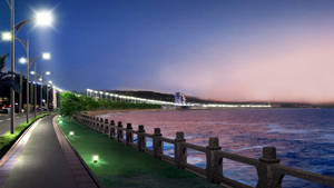 黃昏時分亮燈的海邊步道，遠方有海洋和城市燈光。 — 夜晚 壁紙