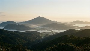 日出籠罩著薄霧的沿海山脈和海灣，有綠色山丘和小鎮。 — 對馬戰鬼 壁紙