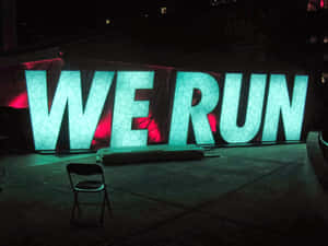 夜間以紅色背光照明的巨大發光青綠色字母WE RUN，附近有一張椅子。 — Running 壁紙