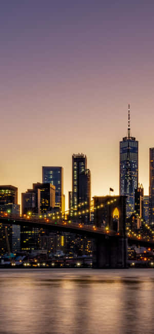 暮色中的布魯克林大橋，城市燈光倒映在水面上。 — iPhone 17e 壁紙