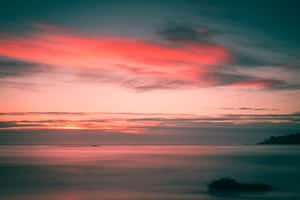 平靜的水面和遠處陸地上空，粉紅色和橙色雲彩映襯下的寧靜日落。 — MacBook Air M3 壁紙