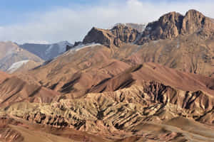 淡藍色天空下，崎嶇的層疊山脈呈現大地色調，帶有雪頂。 — 阿富汗 壁紙