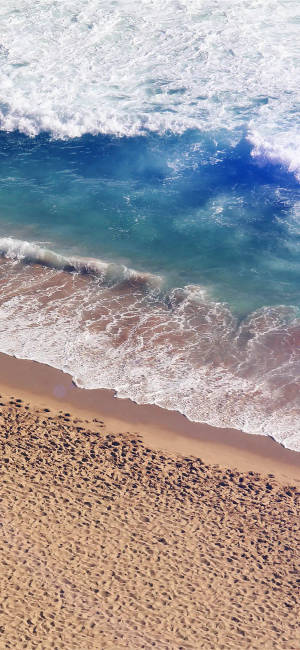 從空中俯瞰，海浪與沙灘交匯，泛起白色泡沫。 — 海灘 iPhone 壁紙