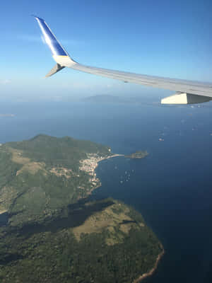 飛機機翼俯瞰熱帶島嶼，有綠色山丘和沿海村莊。 — 3:4 壁紙