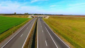 一條高速公路在明亮的藍天下穿過綠色和金色的田野。 — 16:9 壁紙
