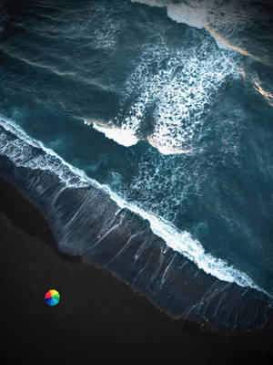 從高空拍攝，海浪拍打黑色沙灘，有一把細小的彩色雨傘。 — 波浪 壁紙