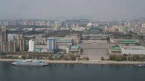 一條寬闊的河流上有一艘船，經過一個建築密集的城市，天空朦朧。 — 韓國 壁紙