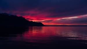 平靜水面上反射出鮮豔的紅色和紫色日落，伴隨著山巒剪影。 — 夕陽天空 壁紙