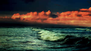 海浪在火紅的日落天空下翻騰，伴隨著烏雲。 — 海灘日落 壁紙