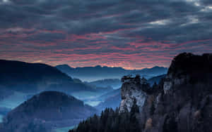 暮色山景，紫色和粉紅色雲彩籠罩著森林覆蓋的山丘。 — 日落 壁紙