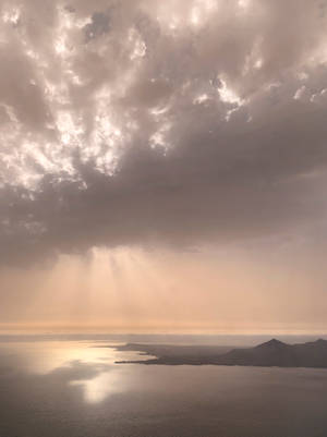 陽光穿過暴風雲，照耀在平靜的海面和遠處的陸地上。 — 頂級 iPhone 壁紙