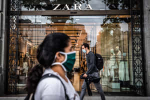 兩名戴口罩的行人走過一間有玻璃窗和裝飾鐵閘的Zara服裝店。 — Zara 壁紙