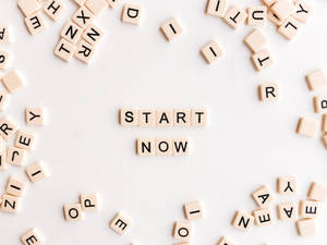 米色字母瓷磚在白色表面上拼出 start now，周圍有散落的字母。 — 激勵 壁紙